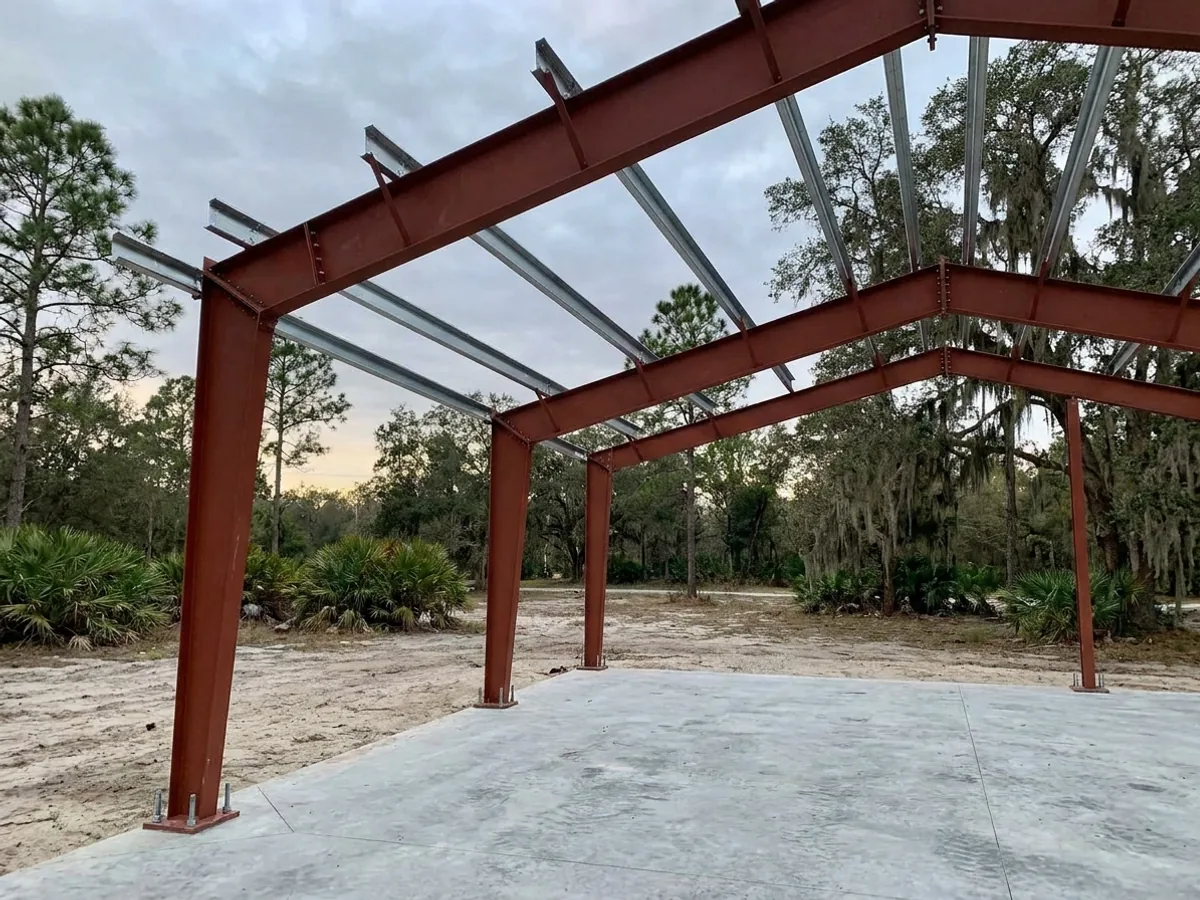 A red oxide primed structural steel frame partly erected on a concrete slab, showing bolted knee joints and anchor bolts.